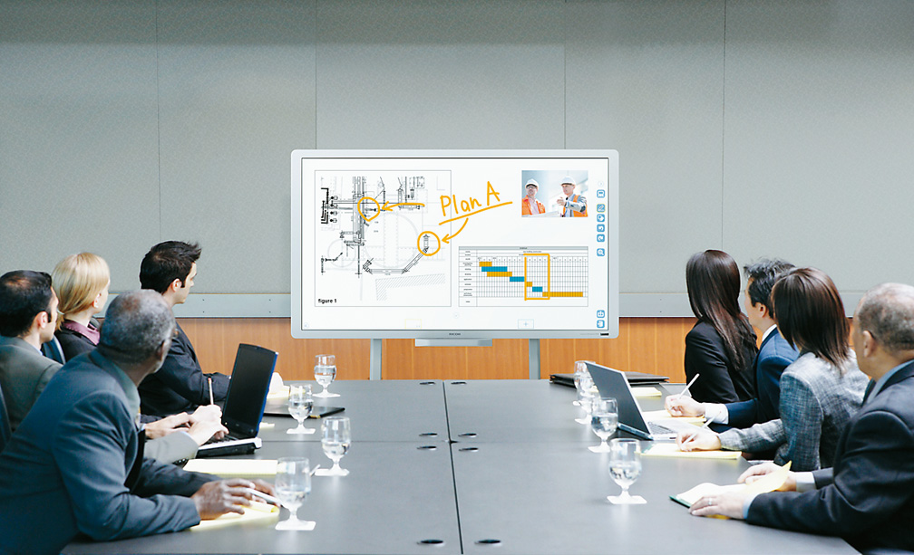 Executives collaborating around a massive Ricoh interactive whiteboard display in a boardroom